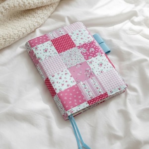 Puede incluir: Una cubierta de diario de patchwork con una variedad de patrones florales y de lunares rosas y celeste. La cubierta tiene un marcador celeste y está sobre una superficie blanca. La cubierta es una mezcla de diferentes telas.