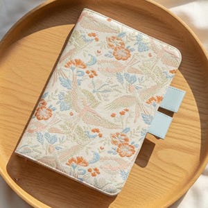 May include: A floral patterned journal cover with a light blue tab is placed on a wooden tray. The cover features a cream background with orange, blue, and pink floral and bird designs. A cream-colored knitted blanket is in the background.