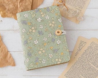 Puede incluir: Una cubierta de diario con estampado floral verde claro con cierre de botón de madera. La cubierta presenta un patrón repetido de flores blancas y amarillas y hojas verdes. El diario se cierra con una cuerda blanca y un botón de madera.