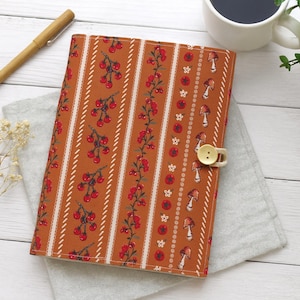 May include: A fabric book cover with a warm orange background and a repeating pattern of cherries, flowers, and mushrooms. The cover is closed with a beige button and loop. A pen and a cup of coffee are in the background.