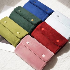 May include: A collection of colorful, rectangular, velvet-like cases with a flap closure and a single button. The cases are in various colors, including green, blue, red, pink, and white. The cases are arranged on a white surface.