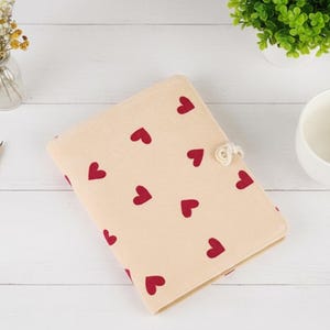 Pode incluir: Uma capa de caderno bege com um padrão de corações vermelhos. A capa tem um fecho de botão branco em forma de flor. Um lápis, uma chávena e pequenas plantas estão no fundo.