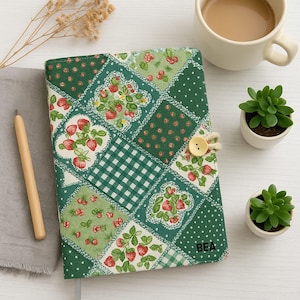 Puede incluir: Una cubierta de diario de patchwork verde y blanco con un estampado de fresas, asegurada con un cierre de botón. Las iniciales "BEA" están en la cubierta. También se ven una taza de café y pequeñas suculentas en macetas.