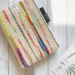 Può includere: Una copertina per notebook beige con strisce verticali di filato colorato nei toni del giallo, verde, rosso e blu. Una tasca in pelle nera è visibile sul lato destro. La copertina è appoggiata su una superficie di legno bianco.