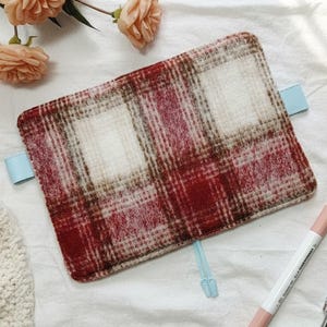 Peut inclure: Une couverture de journal rectangulaire à carreaux dans les tons de rouge, de marron et de crème. La couverture a des languettes bleu clair et est entourée de fleurs et de stylos. Le motif à carreaux est un design classique.