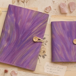 Op de afbeelding: Twee paarse gemarmerde stoffen tijdschriften met houten knoopsluitingen worden gepresenteerd op vintage papier. Gedroogde bloemen en kristallen omringen de tijdschriften. Eén tijdschrift is open en de andere is gesloten. De tijdschriften hebben een uniek, artistiek ontwerp.