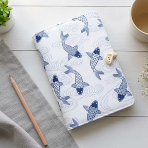 May include: A white notebook cover with a blue koi fish pattern. The cover features a repeating design of stylized fish swimming in a wavy pattern. A beige clasp secures the notebook. A pencil and a gray cloth are in the background.