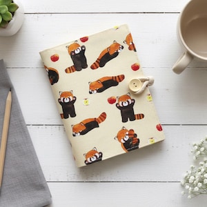 May include: A cream-colored book cover featuring a repeating pattern of red pandas, apples, and small yellow flowers. The cover has a button closure. A pencil and a mug are also visible.