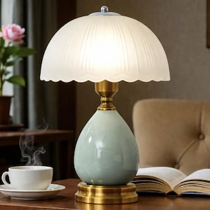 Puede incluir: Una lámpara de mesa de estilo vintage con una pantalla blanca festoneada y una base de cerámica azul claro. La lámpara tiene detalles en latón y un pequeño remate decorativo. También se ven una taza de café y un libro.