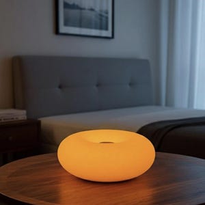 May include: A glowing, donut-shaped lamp with a warm, orange hue sits on a round, wooden table. The lamp's design features a central hole, and it emits a soft light. The background includes a bed and a framed picture.