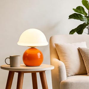 May include: A modern orange and white table lamp with a spherical base and a rounded lampshade. The lamp sits on a small, round, light-colored table next to a neutral-colored mug. A beige armchair and green plant are in the background.