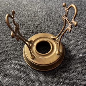 May include: A vintage brass candle holder with a circular base and ornate, curved handles. The candle holder has a central opening for the candle and decorative flourishes on the handles. The brass has a warm, aged patina.