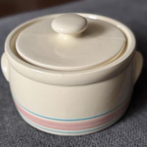 Peut inclure: Plat à gratin en céramique crème avec un couvercle et deux poignées latérales. Le plat présente un motif de rayures roses et bleues autour de la base. Le couvercle a un petit bouton rond.