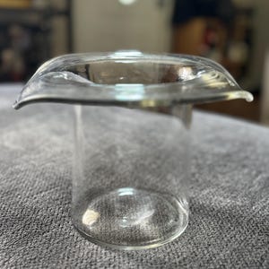 Puede incluir: Un tapón de botella de vidrio transparente con una parte superior ensanchada y redondeada. El tapón tiene un cuerpo cilíndrico y un borde ancho y curvado. El vidrio es transparente, permitiendo el paso de la luz. El tapón está diseñado para encajar en una abertura de botella.