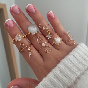Könnte beinhalten: Eine Hand präsentiert eine Sammlung goldfarbener Drahtringe. Die Ringe weisen verschiedene Designs auf, darunter klare und weiße Edelsteine, zarte Wirbel und einen kleinen floralen Anhänger. Die Finger sind mit Maniküre versehen.