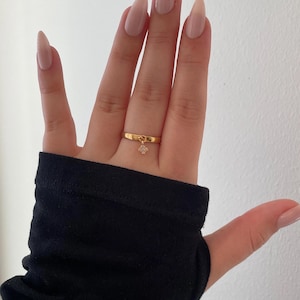 Könnte beinhalten: Ein goldener Ring mit einem kleinen, baumelnden Anhänger mit einem Cluster aus klaren Steinen. Der Ring wird an einer Hand mit langen, mandelförmigen Nägeln getragen, die in einer Nude-Farbe lackiert sind. Der Ärmel eines schwarzen Kleidungsstücks ist sichtbar.