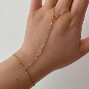 May include: A delicate gold hand chain bracelet. The bracelet features a chain that wraps around the wrist and connects to a ring chain that goes over the middle finger. The jewelry is made of a thin, shiny gold material.