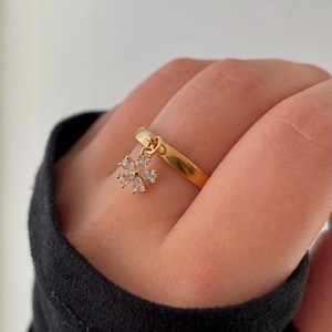 Könnte beinhalten: Ein goldfarbener Ring mit einem kleinen, baumelnden Blumenanhänger. Der Anhänger besteht aus klaren, funkelnden Steinen, die in einem floralen Muster angeordnet sind. Der Ring wird an einem Finger getragen, im Hintergrund ein schwarzer Ärmel.