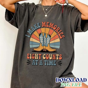 Könnte beinhalten: Ein anthrazitfarbenes Kurzarm-T-Shirt mit einer Grafik aus zwei Cowboystiefeln, zentriert über einem Retro-Sonnenstrahlen-Motiv. Der Text lautet I MAKE MEMORIES EIGHT COUNTS AT A TIME in einer mehrfarbigen, geschwungenen Schriftart.