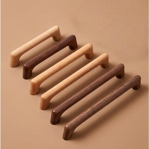 May include: Six wooden cabinet pulls in varying shades of brown. The pulls are rectangular with a curved handle design. The wood tones range from light tan to dark walnut, showcasing the natural grain of the wood. These pulls are ideal for kitchen or bathroom cabinets.