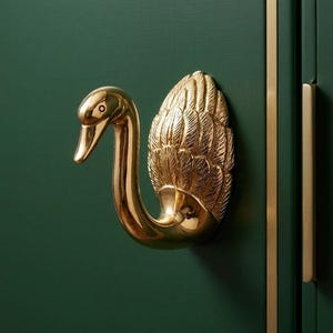 May include: A decorative cabinet pull in the shape of a swan, finished in gold. The swan's head and neck are curved, and the body features detailed feathering. It's mounted on a dark green cabinet door, with gold trim and a matching handle.