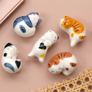 May include: Five ceramic cat figurines in various poses and colors, including white with blue, orange, and brown markings. Each cat is curled up or lying down, with painted details. The figurines are on a pink surface.