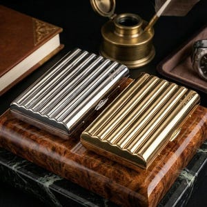 Puede incluir: Dos pitilleras metálicas, una plateada y otra dorada, con diseño acanalado. Las pitilleras son rectangulares con borde ondulado y un pequeño cierre. Las pitilleras están sobre una superficie de madera con base de mármol.