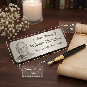 Puede incluir: Placa conmemorativa plateada con la inscripción "In Loving Memory Of William Thompson" y las fechas "August 15, 1946 - April 8, 2024". La placa presenta un retrato y espacio para nombres y fechas personalizados. Un bolígrafo negro y dorado está cerca.