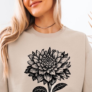 May include: A beige sweatshirt with a large black and white floral design. The floral graphic is centered on the front of the sweatshirt and includes a detailed depiction of petals, leaves, and stem. The sweatshirt is paired with light blue jeans.