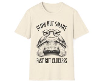 May include: Cream-colored t-shirt featuring a black and white illustration of a turtle wearing glasses. The text on the shirt reads "SLOW BUT SMART" above the turtle and "FAST BUT CLUELESS" below.