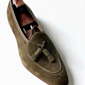 May include: Olive green suede loafer with a tassel detail. The shoe features a rounded toe and a leather sole. A wooden shoe tree is inserted inside the shoe to maintain its shape.