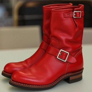 Könnte beinhalten: Ein Paar wadenhohe Stiefel aus leuchtend rotem Leder mit abgerundeter Zehenpartie, einem Absatz in Schichtholzoptik und zwei silberfarbenen Metallschnallen. Eine Schnalle befindet sich über dem Spann, die andere am oberen Schaftrand.