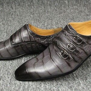 K&ouml;nnte beinhalten: Ein Paar graue Lederschuhe mit Krokodilhautstruktur. Die Schuhe haben eine spitze Zehenpartie und drei silberne Schnallen. Das Innenfutter ist hellbraun. Die Schuhe werden auf einer grauen Oberfl&auml;che pr&auml;sentiert.