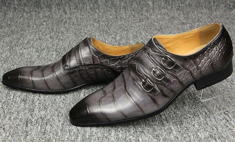 K&ouml;nnte beinhalten: Ein Paar graue Lederschuhe mit Krokodilhautstruktur. Die Schuhe haben eine spitze Zehenpartie und drei silberne Schnallen. Das Innenfutter ist hellbraun. Die Schuhe werden auf einer grauen Oberfl&auml;che pr&auml;sentiert.