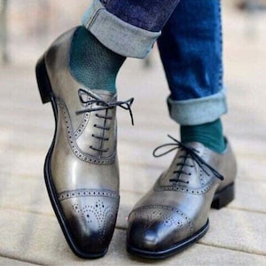 May include: A pair of grey leather dress shoes with black laces and a cap toe design. The shoes feature decorative perforations and are worn with blue jeans and green socks. The shoes have a classic, formal style.