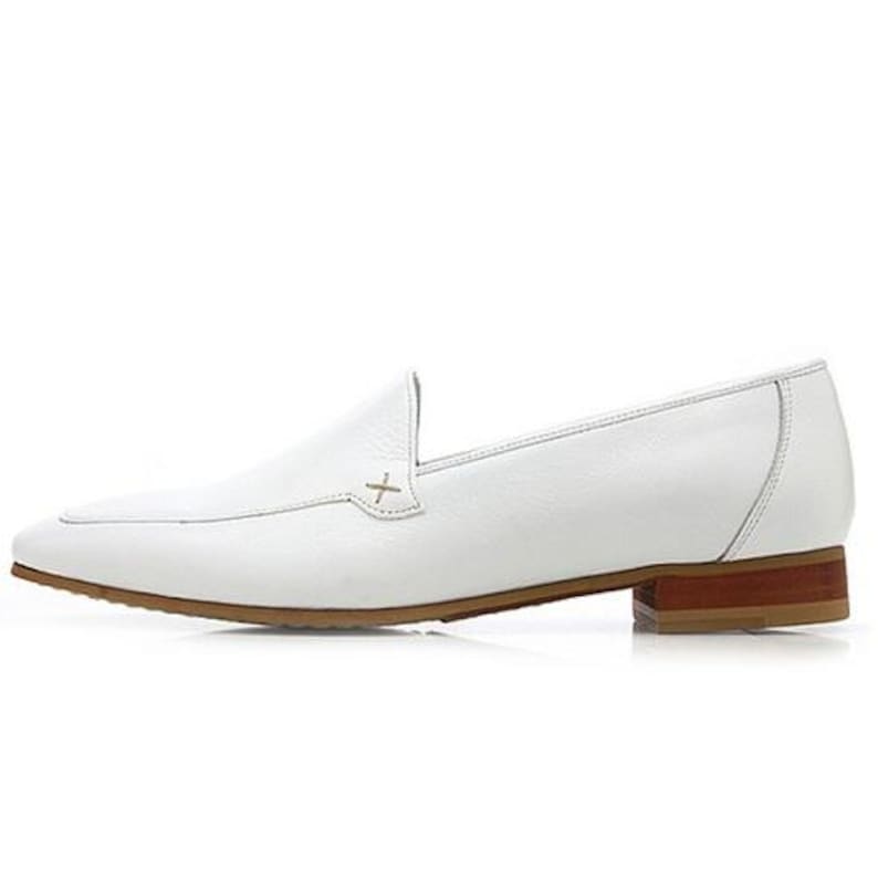 May include: A white leather loafer with a brown sole and heel. The shoe features a pointed toe and a decorative cross-stitch detail on the vamp. The heel measures approximately 1 inch.
