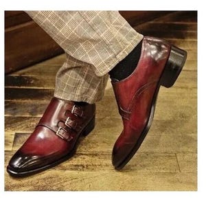 May include: Burgundy leather dress shoes with a polished finish. One shoe features a double monk strap with three buckles, the other a minimalist design. The shoes are worn with plaid trousers.