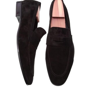 May include: A pair of black suede loafers with wooden shoe trees inside. The shoes have a classic slip-on design with a stitched sole and a small heel. The suede has a soft, matte finish.