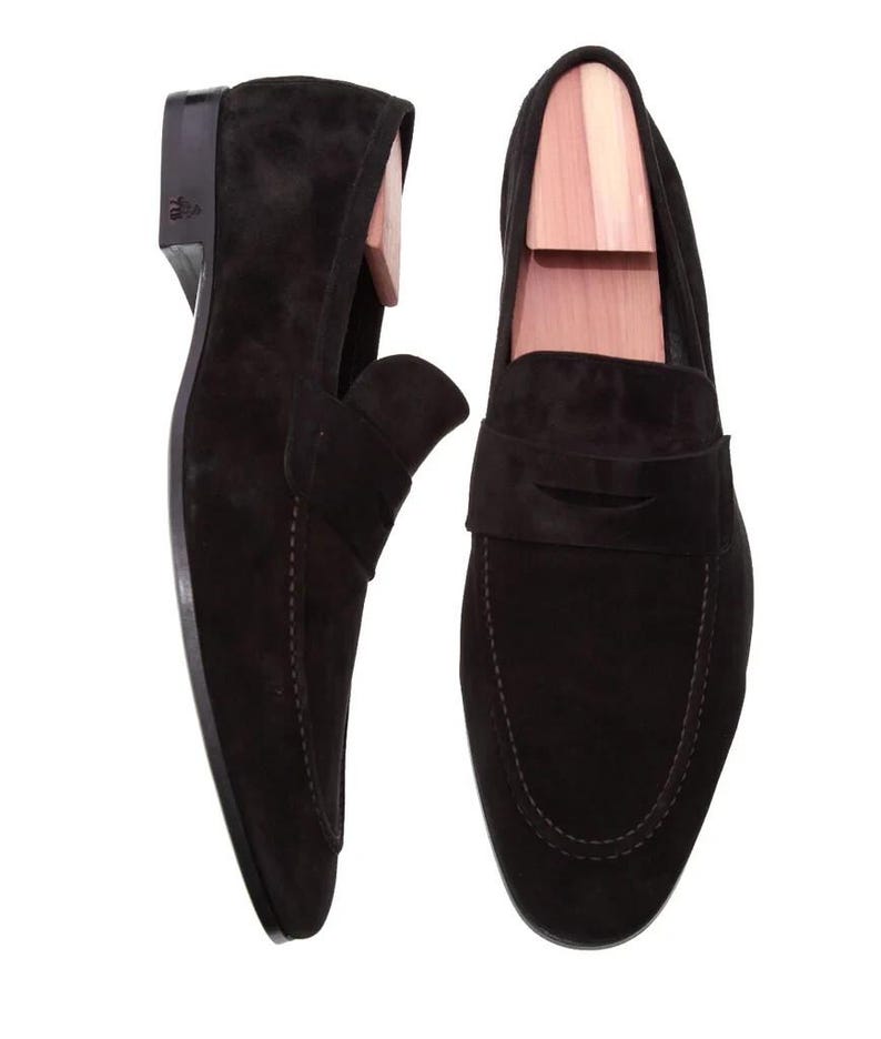 May include: A pair of black suede loafers with wooden shoe trees inside. The shoes have a classic slip-on design with a stitched sole and a small heel. The suede has a soft, matte finish.