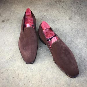 May include: A pair of brown suede slip-on shoes with wooden shoe trees inside. The shoes have a rounded toe and a dark brown sole. The shoe trees are a reddish-pink color. The shoes are displayed on a gray surface.