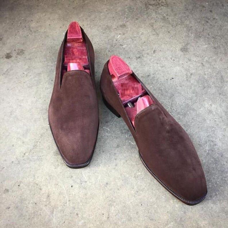 May include: A pair of brown suede slip-on shoes with wooden shoe trees inside. The shoes have a rounded toe and a dark brown sole. The shoe trees are a reddish-pink color. The shoes are displayed on a gray surface.