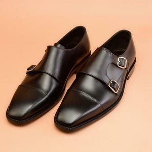 May include: A pair of black leather dress shoes with double monk straps and gold-colored buckles. The shoes have a square toe design and are set against a peach-colored background.