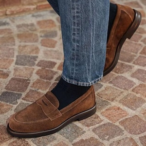 May include: Brown suede penny loafers with a dark brown sole. The shoes are worn with blue jeans and navy socks. The loafers have a classic design with a strap across the top.