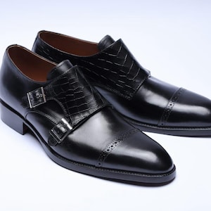 May include: A pair of black leather dress shoes with a glossy finish. The shoes feature a double monk strap design with silver buckles and textured accents on the straps. The toe caps have decorative perforations.