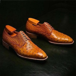 May include: A pair of brown leather dress shoes with a textured ostrich pattern. The shoes have a classic lace-up design, a low heel, and wooden shoe trees inside. The shoes are displayed on a dark green surface.