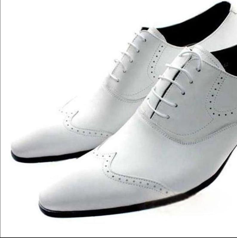 May include: A pair of white leather dress shoes with a classic Oxford design. The shoes feature a lace-up closure, decorative perforations, and a black sole. The shoes are designed for formal occasions.