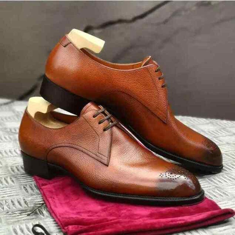Op de afbeelding: Een paar bruine leren herenschoenen met een klassiek ontwerp. De schoenen hebben een gepolijste afwerking, een vetersluiting en een kleine hak. Ze worden gepresenteerd op een rode doek.