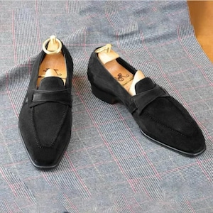 Pode incluir: Um par de mocassins pretos de camur&ccedil;a com um design cl&aacute;ssico. Os sapatos apresentam uma biqueira elegante e uma tira na parte superior. As formas s&atilde;o inseridas no interior dos sapatos. Os mocassins s&atilde;o exibidos sobre um tecido xadrez cinza.