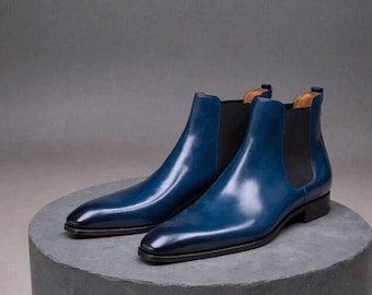May include: A pair of blue Chelsea boots with a glossy finish. The boots have a sleek design with elastic side panels and a low heel. The boots are displayed on a grey, circular platform.