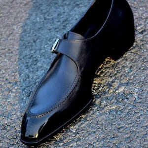 May include: A black leather dress shoe with a pointed toe and a strap with a silver buckle. The shoe has a glossy finish and is set against a textured surface.